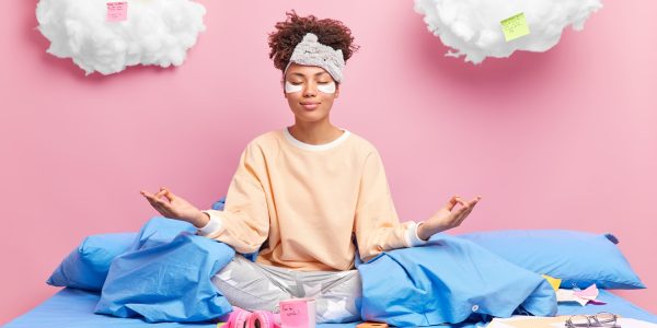 Pleased African American woman meditates in bed feels relaxed wears nightwear closes eyes practices yoga after studying drinks coffee isolated over pink background. Distance studying concept