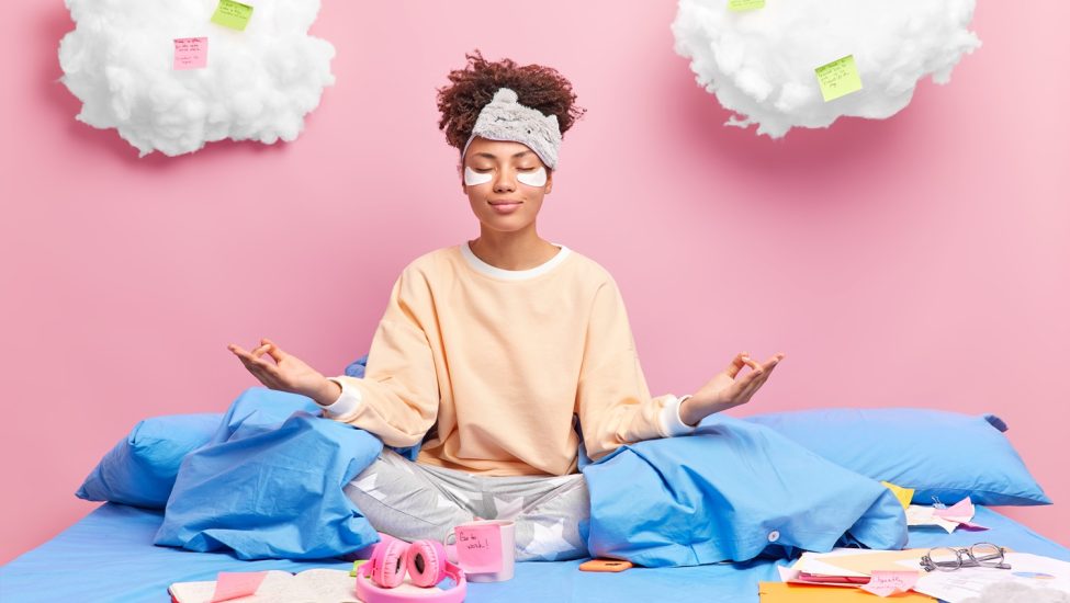 Pleased African American woman meditates in bed feels relaxed wears nightwear closes eyes practices yoga after studying drinks coffee isolated over pink background. Distance studying concept